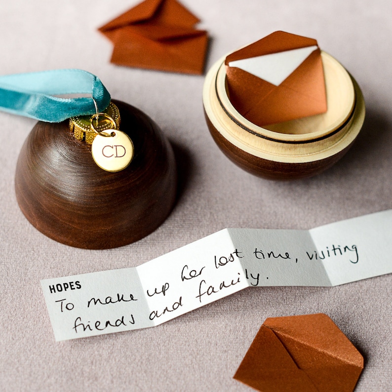 May include: A wooden ornament shaped like a Christmas tree bauble with a gold-colored metal charm that says "CD" on it. The ornament is sitting on a light pink surface next to a small wooden box with a brown envelope inside. A folded piece of paper with the text "HOPES To make up her lost time, visiting friends and family." is also on the surface.