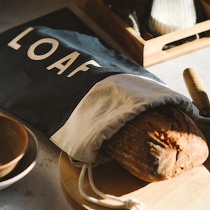 May include: A black and white canvas bread bag with the word "LOAF" printed in white. The bag is filled with a loaf of sourdough bread and is sitting on a wooden chopping board.