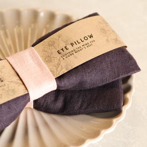 May include: A dark purple linen eye pillow with a brown paper label that reads "Eye Pillow - Soothing the mind for a good night's rest".