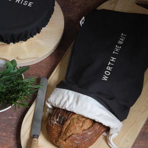 May include: A black bread bag with white text reading "WORTH THE WAIT", containing a loaf of bread. A round black cover with "EAT & RISE" is also present. The items are on a wooden board.