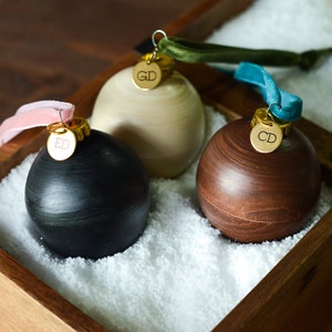 May include: Three wooden ornaments with personalized gold tags. The ornaments are in a wooden box filled with white faux snow. The tags read "GD", "ED", and "CD".
