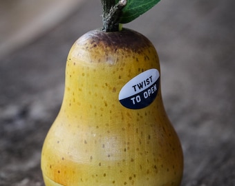 May include: A wooden pear-shaped container with a yellow body and a brown top. It has a green leaf and a twig stem. A sticker on the front reads "TWIST TO OPEN".