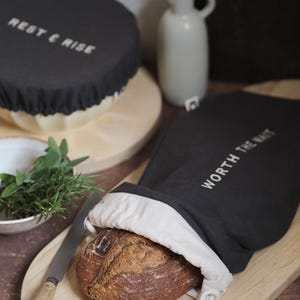 Könnte beinhalten: Eine dunkelgraue Brottasche mit dem Text "WORTH THE WAIT" und eine runde Brotdecke mit "REST & RISE" in weißen Buchstaben. Ein Brotlaib liegt in der Tasche auf einem Holzschneidebrett. Ein Messer und Kräuter sind ebenfalls vorhanden.