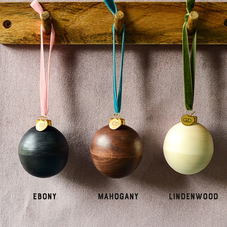 May include: Three wooden Christmas ornaments hanging from hooks on a wooden shelf. The ornaments are made of ebony, mahogany, and lindenwood. Each ornament has a gold tag with a letter abbreviation.