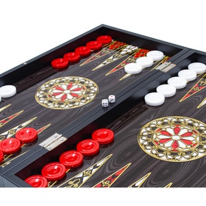 Könnte beinhalten: Ein schwarzes Backgammon-Set aus Holz mit roten und weißen Spielsteinen. Das Brett ist geöffnet und zeigt die Spielfläche mit einem dekorativen Design. Zwei Würfel liegen auf dem Brett.
