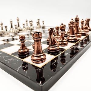 Op de afbeelding: Een zwart-wit marmeren schaakbord met koperen schaakstukken. Het bord heeft een gouden rand en de stukken zijn in de beginpositie gerangschikt.