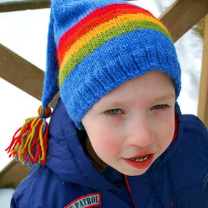 Puede incluir: Un niño lleva un gorro azul tejido con un patrón de rayas de arcoíris y una chaqueta azul con un parche de "White Patrol". El gorro tiene una borla larga hecha de hilo de colores.