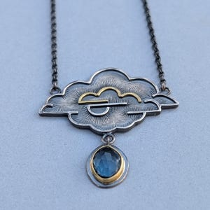 Può includere: Una collana in argento e oro con un ciondolo a forma di nuvola e una goccia di pietra preziosa blu. La nuvola ha una superficie testurizzata e un design a forma di sole dorato.