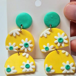 Puede incluir: Un par de pendientes colgantes con un poste circular de color verde azulado conectado a dos dijes de semicírculo amarillos apilados. Cada pieza amarilla está decorada con flores blancas con centros verde azulado, unidas por anillas de metal dorado.