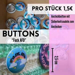 Könnte beinhalten: Eine Auswahl runder Buttons mit Sicherheitsnadeln. Die Buttons sind hellblau mit einem Regenbogen und dem Text "FUCK AFD". Das Wort "BUTTONS" ist in großen schwarzen Buchstaben dargestellt. Die Buttons haben einen Durchmesser von ca. 2,5 cm.