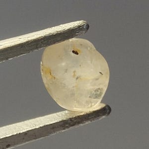 May include: A small, translucent, pale yellow gemstone with an irregular, rounded shape. The stone features visible internal inclusions and natural fractures, held securely by a pair of metal tweezers with a textured, silver-toned finish.