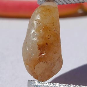 Peut inclure: Une petite pierre translucide de forme irrégulière avec une surface lisse et polie. La pierre présente une palette de couleurs chaudes et marbrées de beige pâle, de crème et de taches orange ambré, tenue verticalement par une pince en métal argenté.