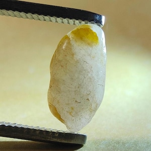 May include: A small, irregularly shaped, translucent white gemstone with a smooth, polished surface and a distinct patch of vibrant yellow color near the top edge, held securely between the metal tips of a pair of silver-toned tweezers.
