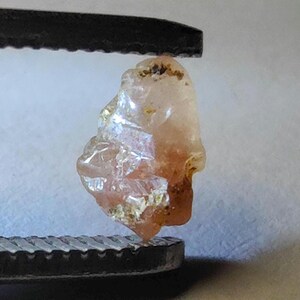 May include: A raw, translucent peach-colored gemstone held by black metal tweezers. The stone features an irregular, multifaceted surface with visible internal inclusions and light-reflecting crystalline structures throughout its body.