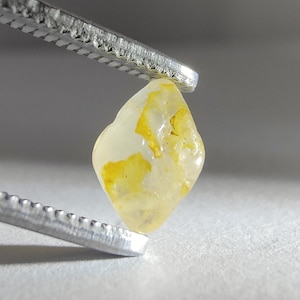May include: A small, translucent, pale yellow gemstone with irregular, vibrant yellow inclusions held by a pair of silver-toned metal tweezers. The stone has a smooth, rounded, diamond-like shape with a polished surface that reflects light.