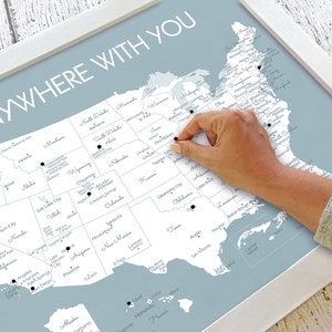May include: A framed map of the United States with the words "ANYWHERE WITH YOU" in white. The map is light blue with black dots marking locations. A hand is placing a black pin on the map.