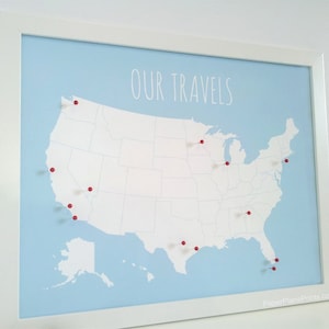 May include: Framed map print of the United States with red push pins marking locations. The map is light blue with the words "OUR TRAVELS" in white script. The frame is white.