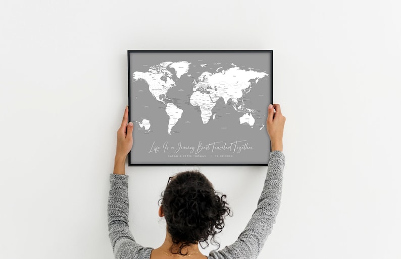 May include: A person hangs a grey world map with white countries and a black frame on a white wall. The map has the text "Let's Go on a Journey Best Travelled Together" with a date.