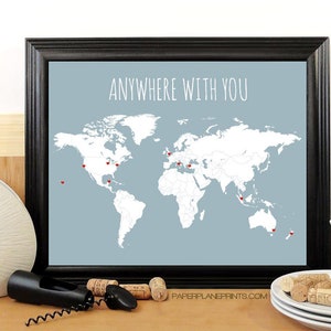 May include: A framed print with a world map on a light blue background. The map is white with red hearts marking locations. The text "Anywhere with you" is printed in white at the top of the map. The text "paperplaneprints.com" is printed in black at the bottom of the map.