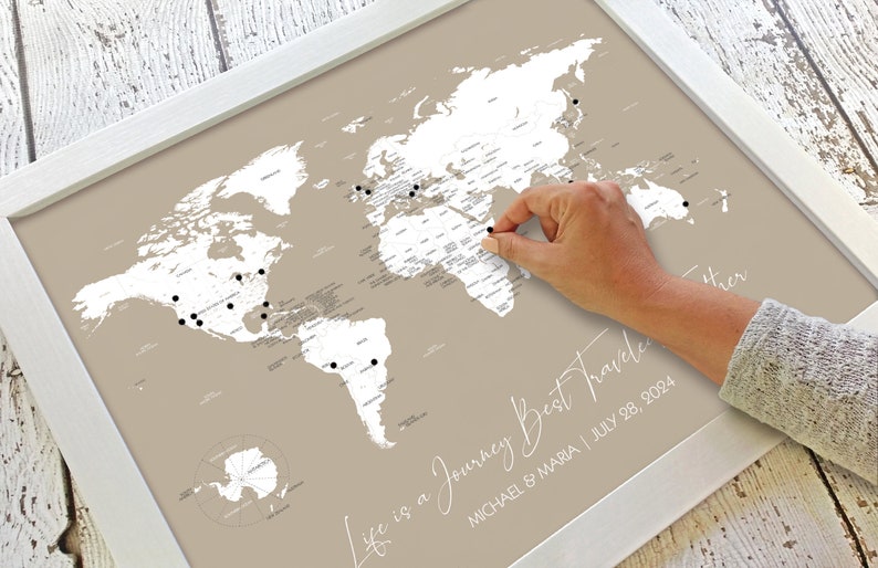 May include: A beige world map with a white frame. The map is designed for marking off places visited with push pins. The text "Life is a Journey Best Traveled" is written in white script at the bottom of the map. The text "MICHAEL & MARIA | JULY 28, 2024" is written in white script below the text "Life is a Journey Best Traveled".
