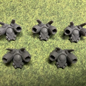 May include: Five identical gray plastic miniature jetpack components, each featuring a central circular housing, two side-mounted thruster nozzles, and two small upward-pointing fins, arranged in a grid pattern on a textured green surface.