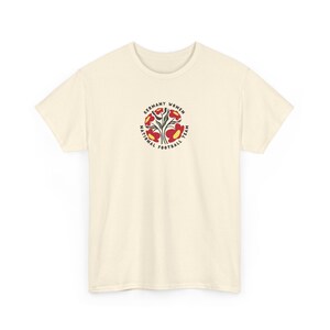 Op de afbeelding: Crèmekleurig T-shirt met een cirkelvormig ontwerp met de tekst "GERMANY WOMEN NATIONAL FOOTBALL TEAM". Het ontwerp bevat gestileerde rode en gele bloemen en bladeren. Het shirt heeft korte mouwen en een ronde hals.