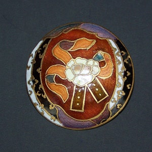 May include: A round, gold-toned button with a colourful enamel design. The design features a white flower with orange and brown ribbons, surrounded by a black and white border.