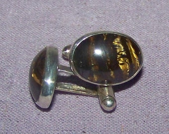 May include: A pair of silver cufflinks with oval-shaped tiger's eye stones. The stones have a dark brown and gold color.