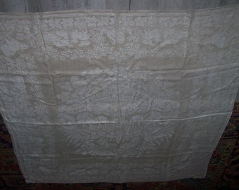 May include: A white linen tablecloth with an intricate embroidered design of a floral pattern. The tablecloth is slightly wrinkled and has a subtle sheen.