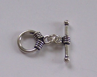 May include: A silver-toned toggle clasp for jewelry making, consisting of a circular ring and a T-bar component. Both pieces feature a decorative ribbed texture with dark recessed lines, connected by two small silver-toned jump rings.