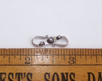 May include: A silver-toned metal S-hook clasp featuring decorative filigree-style detailing and small spherical accents. The clasp is positioned above a wooden ruler, showing a length of approximately 1.25 inches from end to end.