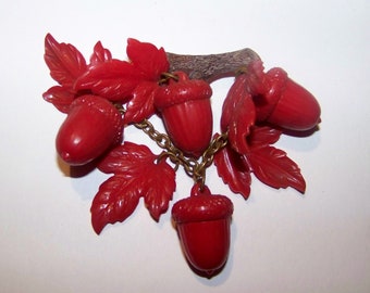 May include: A red plastic brooch in the shape of an acorn branch with leaves and acorns. The branch is brown and the acorns are connected by a gold chain.