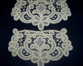 May include: Two off-white lace doilies with intricate floral and geometric patterns. Each doily features a central floral motif surrounded by delicate openwork and scalloped edges. The lace is finely detailed, creating a textured appearance against a dark background.