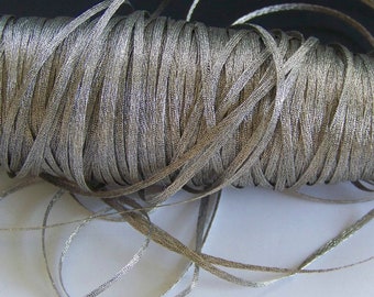 May include: A spool of metallic silver ribbon. The ribbon is tightly wound, revealing a textured surface. The ribbon is thin and flat, with a slight sheen, suitable for crafts and embellishments.