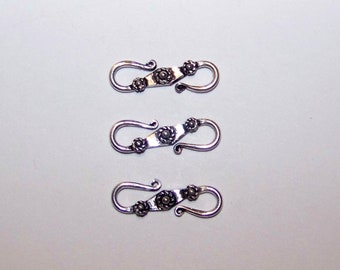 May include: Three silver-toned metal S-hook clasps arranged vertically. Each clasp features a central elongated bar with a decorative floral pattern and a small raised dot in the middle, flanked by two smaller floral motifs, ending in curved hooks.