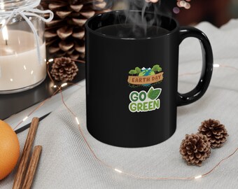 May include: Black coffee mug with a white design that says "Earth Day Go Green" with a green globe and leaves. The mug is sitting on a light brown surface with a string of lights.