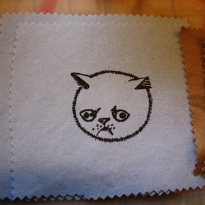 May include: A blue fabric square with a black and white illustration of a cat's face with a sad expression.