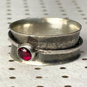 May include: A silver-toned ring with a textured band and a rotating outer ring. A small, round, red gemstone is set on the side. The ring is displayed on a white surface with a dotted pattern. The text "EG Studio Jewelry" is visible.