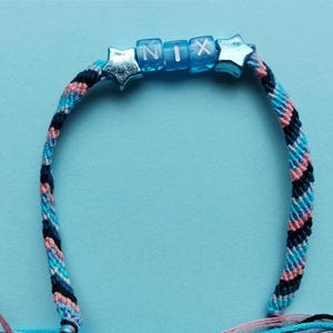 May include: A handmade friendship bracelet in shades of blue, pink, and black. The bracelet features the name "NIX" spelled out with blue letter beads and star-shaped accents. The background is a solid light blue.