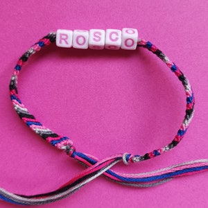 Könnte beinhalten: Ein mehrfarbiges Freundschaftsarmband mit dem Namen "ROSCO" in rosa Buchstaben auf weißen Perlen.