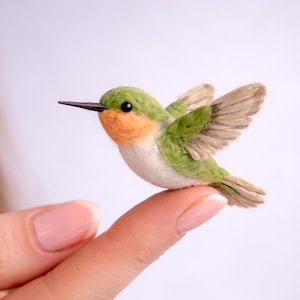 以下が含まれることがあります： 緑、オレンジ、白の色をした手作りのハチドリの置物。鳥は指の上に止まり、翼を広げています。ハチドリは長く黒い嘴を持ち、フェルトで作られています。