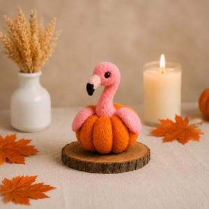 Può includere: Una figura di fenicottero rosa in lana infeltrita con becco dalla punta nera, fissata sopra una zucca arancione rotonda e testurizzata. La figura è posizionata su una base circolare di legno, mostrando una consistenza morbida e soffice.
