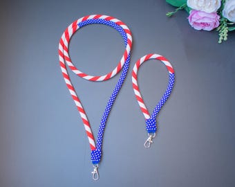 Op de afbeelding: Twee kralen lanyards in de kleuren rood, wit en blauw. De langere lanyard heeft een wit en blauw patroon, terwijl de kortere lanyard een rood en wit patroon heeft. Beide lanyards hebben zilveren sluitingen.