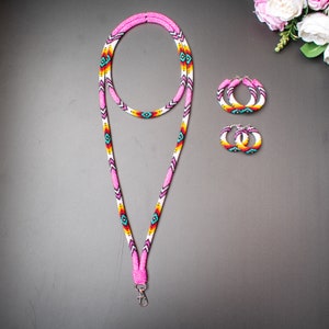 Op de afbeelding: Een roze kralenkoord met een geometrisch patroon in wit, geel, rood en blauw. Het koord heeft een zilveren sluiting. Twee bijpassende roze kralen-hoep-oorbellen met hetzelfde geometrische patroon zijn ook inbegrepen.
