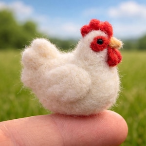 Può includere: Un piccolo pollo fatto a mano in feltro bianco con cresta rossa, bargigli e occhio nero. Il pollo ha un becco giallo ed è posato su un dito. Lo sfondo è un campo verde sfocato e un cielo blu.