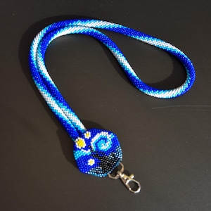 May include: A blue and white beaded lanyard with a blue, white, and yellow beaded charm. The charm has a flower design.
