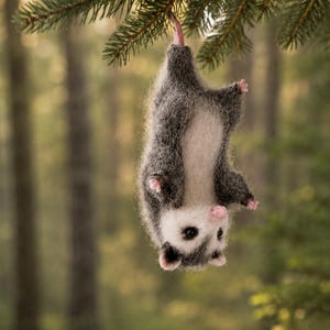 Könnte beinhalten: Eine aus Nadelfilz gefertigte Opossum-Figur mit dunkelgrauem und weißem Fell sowie rosa Nase, Ohren und Pfoten, die kopfüber an ihrem Schwanz an einem Kiefernzweig hängt. Das Tier hat kleine schwarze Augen und ein flauschiges, strukturiertes Fell.