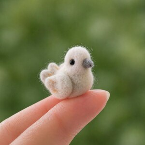 Puede incluir: Un pequeño pájaro de fieltro hecho a mano, en tonos blanco y crema. El pájaro tiene un cuerpo redondeado, pequeñas alas y un pico gris. Está posado en un dedo sobre un fondo verde borroso. El artículo artesanal es una representación en miniatura de una paloma.