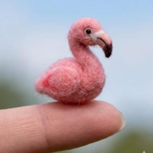 May include: A small, pink flamingo ornament with a black beak and a red marking on its side. The flamingo is crafted from a soft, textured material and is resting on a finger. The background is a blurred blue and green.
