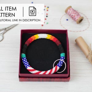 May include: A beaded necklace with a patriotic design, featuring red, white, and blue beads, is displayed in a red box. The necklace has a circular shape and a silver ring. The image also includes scissors, thread, and a scroll.